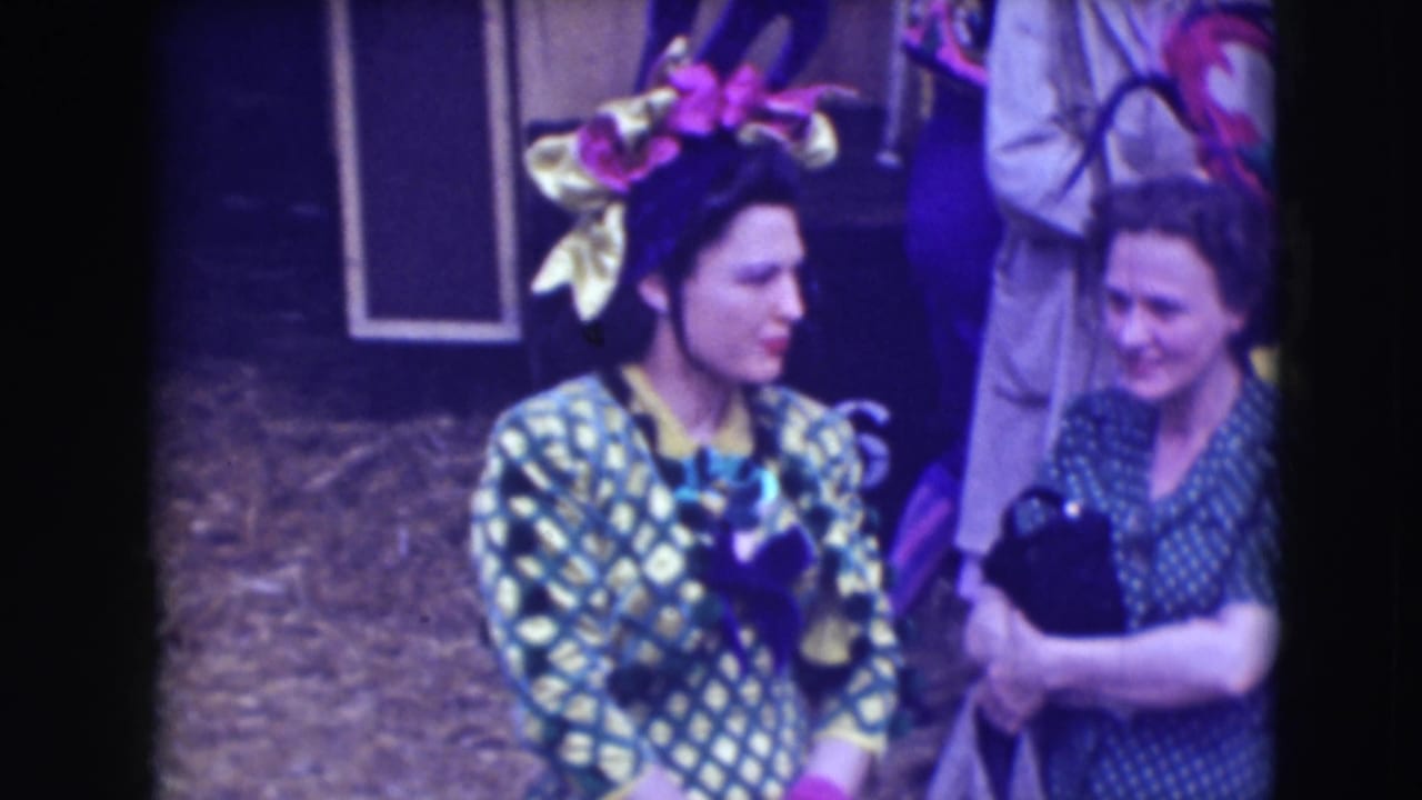 Two women in patterned performance clothing stand near a circus setting with a large floral headpiece visible.