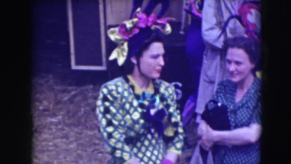 In 1948 Sarasota circus footage, a woman in a green patterned costume and large pink-and-gold headpiece stands beside another woman holding an object.