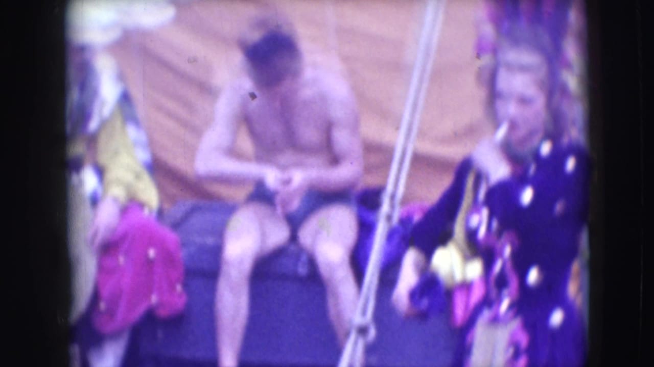 A shirtless male performer sits in the background of the 1948 Sarasota circus scene while costumed figures and props surround him.