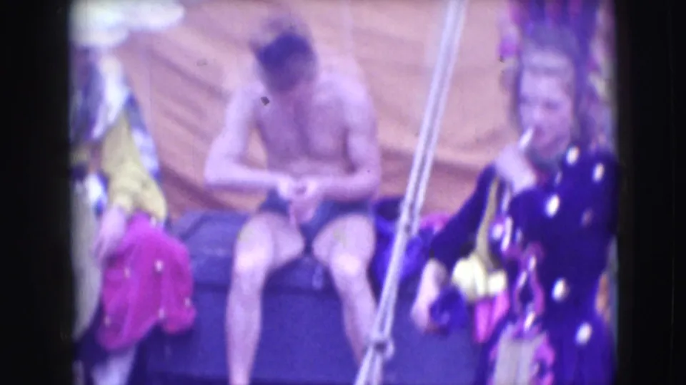 A shirtless male performer sits in the background of the 1948 Sarasota circus scene while costumed figures and props surround him.