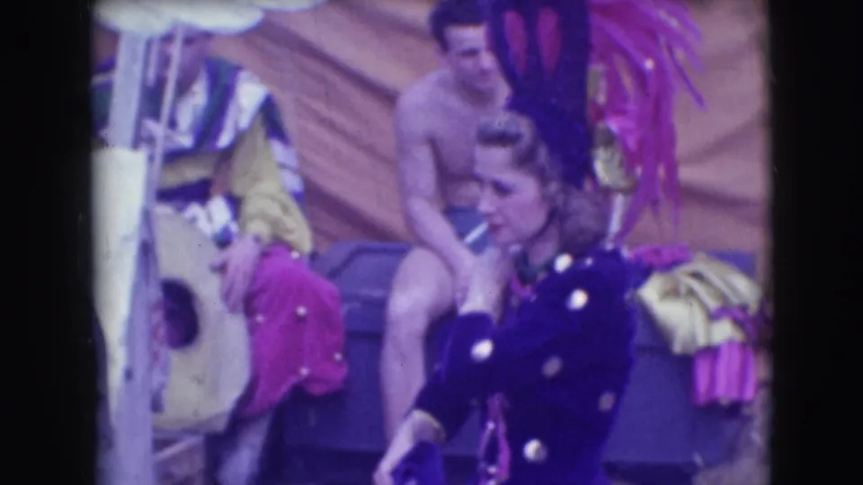 A woman in a dark costume with light spots moves through the 1948 Sarasota circus frame as a seated male performer remains behind her.
