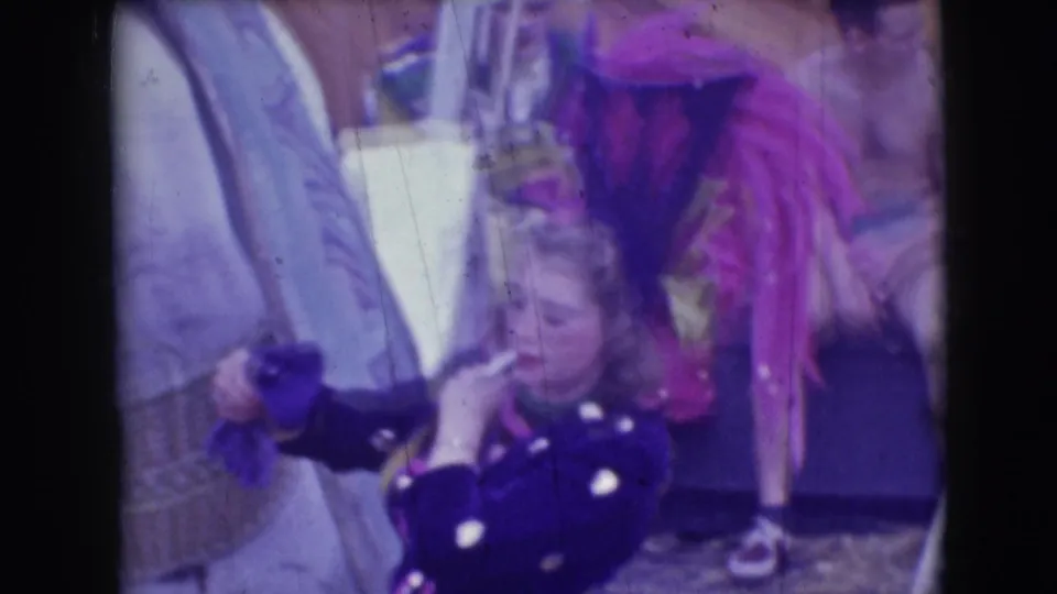 In the Sarasota circus footage, a costumed performer holds a trumpet near pink feathered costume elements and tent equipment.