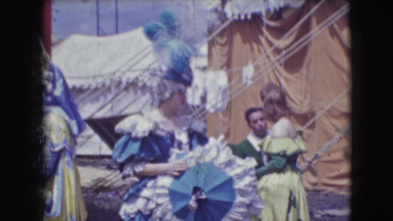 1948 Ringling Brothers Sarasota footage showing women in colorful feathered costumes near tents, with a suited man in the background.