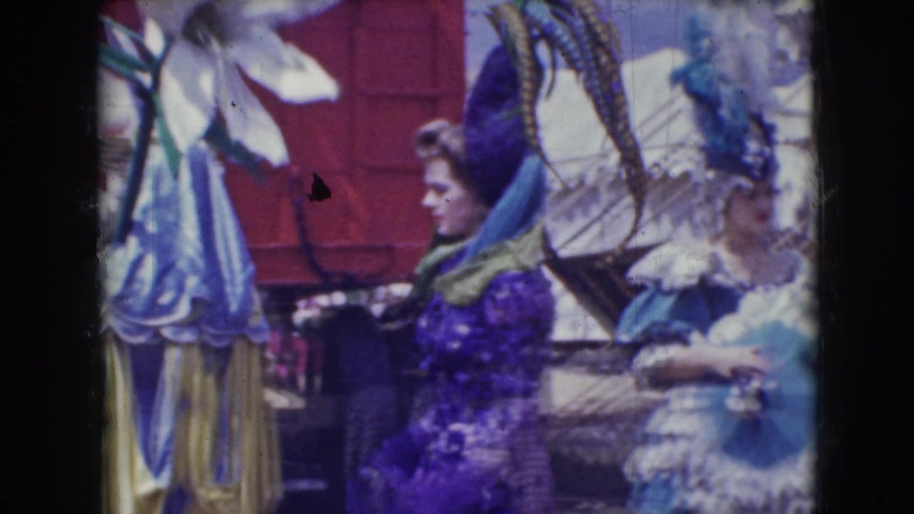 Women in purple, blue, and yellow costumes walk past red panels and circus tenting.