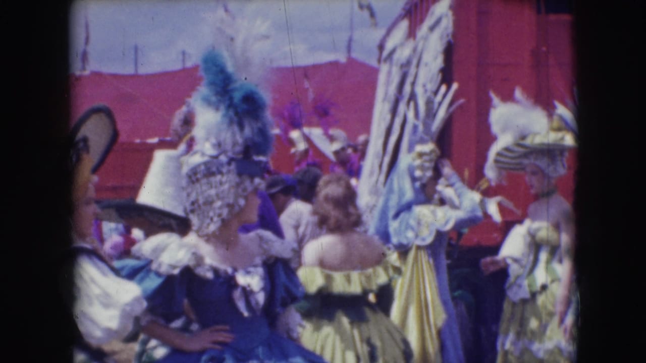 Costumed women with feathered headpieces stand in front of a red circus wagon.