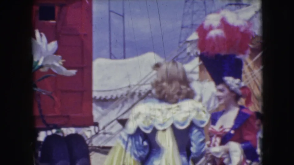 A visitor-shot 1948 circus frame shows ruffled period costumes, feathered hats, a white tent, and red wagon structures.
