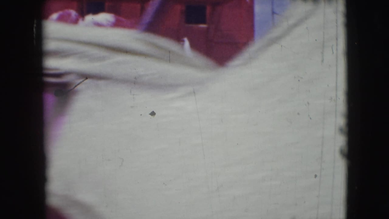 Soft 8mm frame from the Sarasota clip with pale costume fabric close to the lens, dark gate edges and a red backdrop behind it.