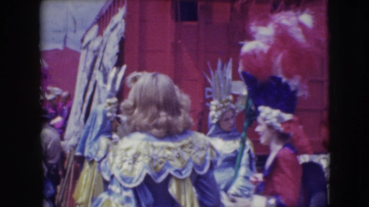 Costumed people with feathered headpieces and ruffled garments stand near a red backdrop.