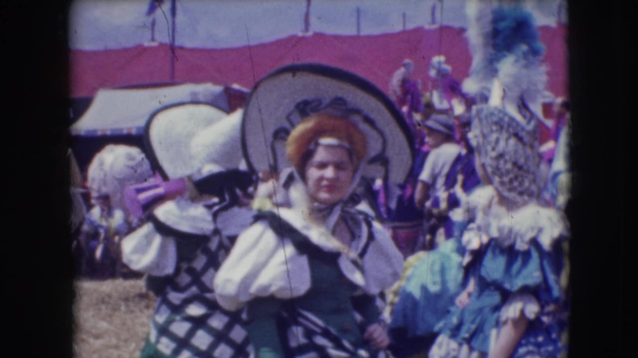 A costumed woman in a broad hat stands among other performers and spectators outdoors.