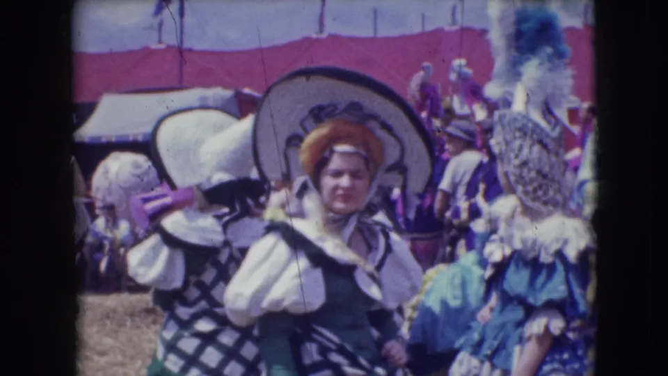 A costumed participant in a broad hat and ruffled clothing stands amid a busy Ringling Brothers outdoor scene in Sarasota.