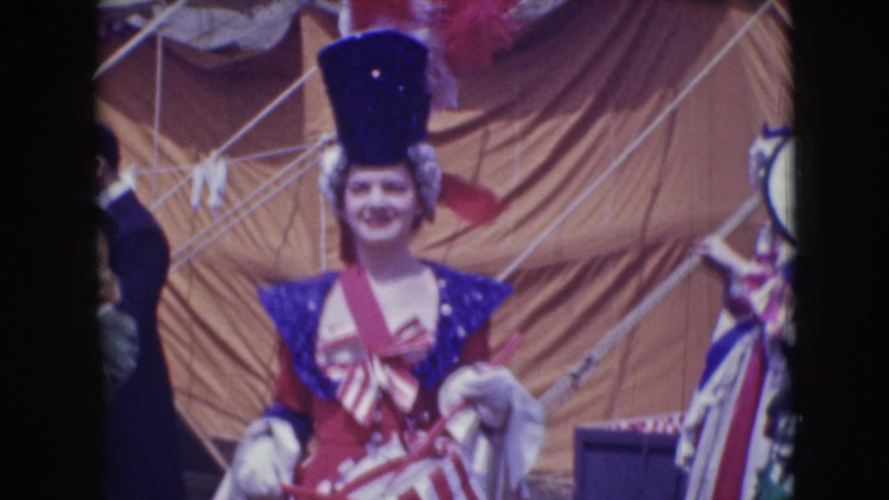 Costumed woman in a tall blue hat stands near tent rigging and other performers.
