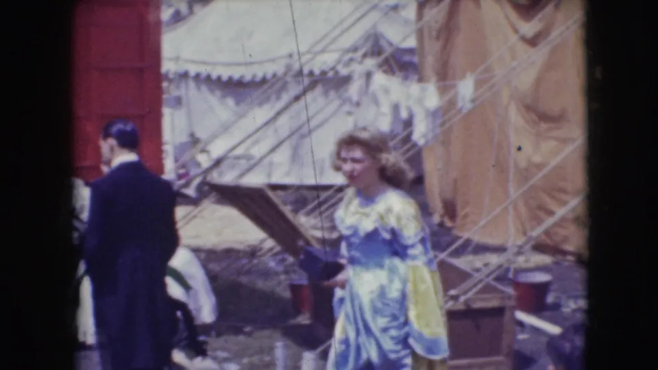 Outside the 1948 Sarasota circus tent area, a suited man and a woman in a pale costume move past canvas walls, ropes, and equipment.