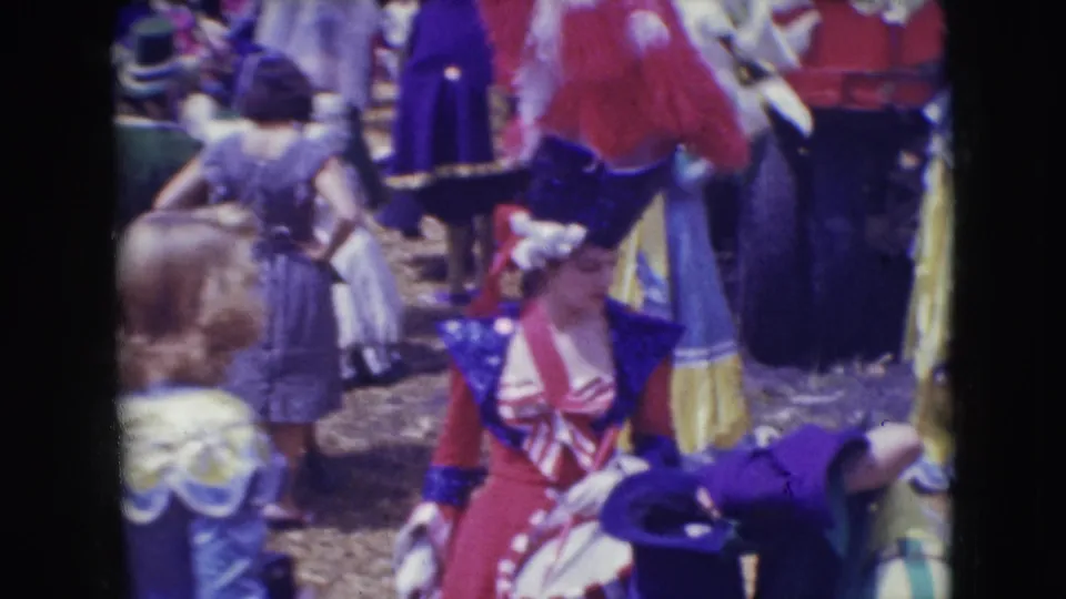 At the Sarasota circus, a performer with a large red feathered headpiece stands near a red wagon and nearby costumed performers.