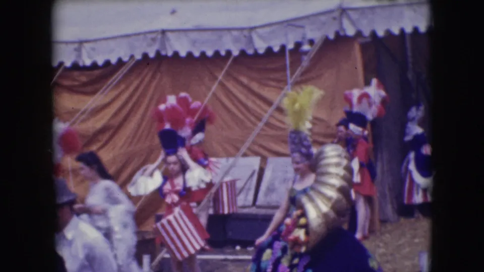 Visitor-shot 1948 Sarasota circus view with a white-costumed performer, feathered figures, flags, and tent canvas.