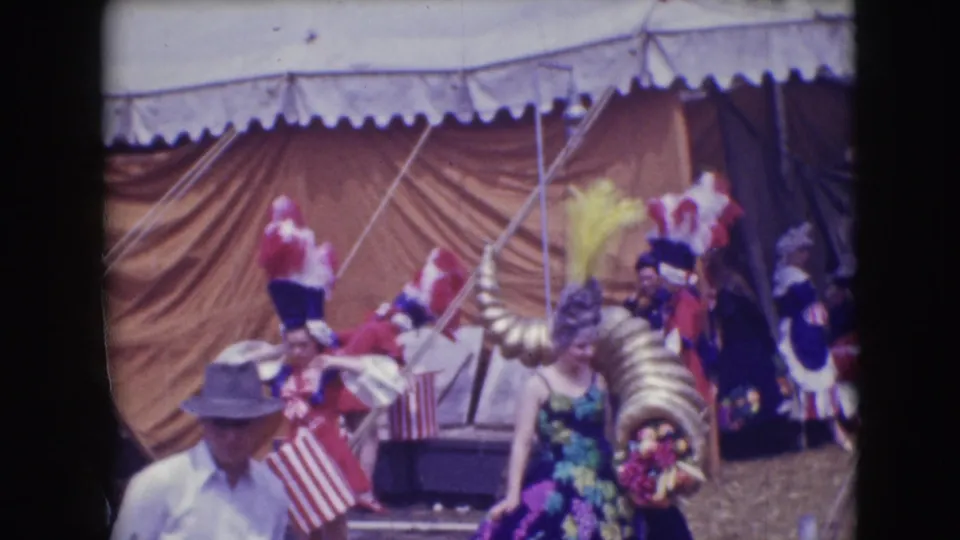 1948 Sarasota circus frame with floral and feathered costumes, a gold horn-shaped prop, tent canvas, and a nearby spectator.