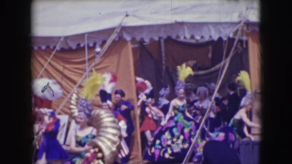 Backstage circus performers in ornate costumes and feathered headwear cluster near tent ropes in the 1948 Sarasota footage.
