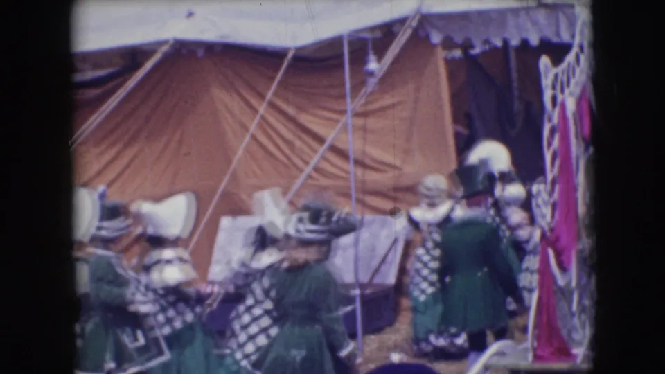 1948 Sarasota circus parade frame with green and white checkered walkers, tall hats, tent rigging, and striped fabric at frame edge.