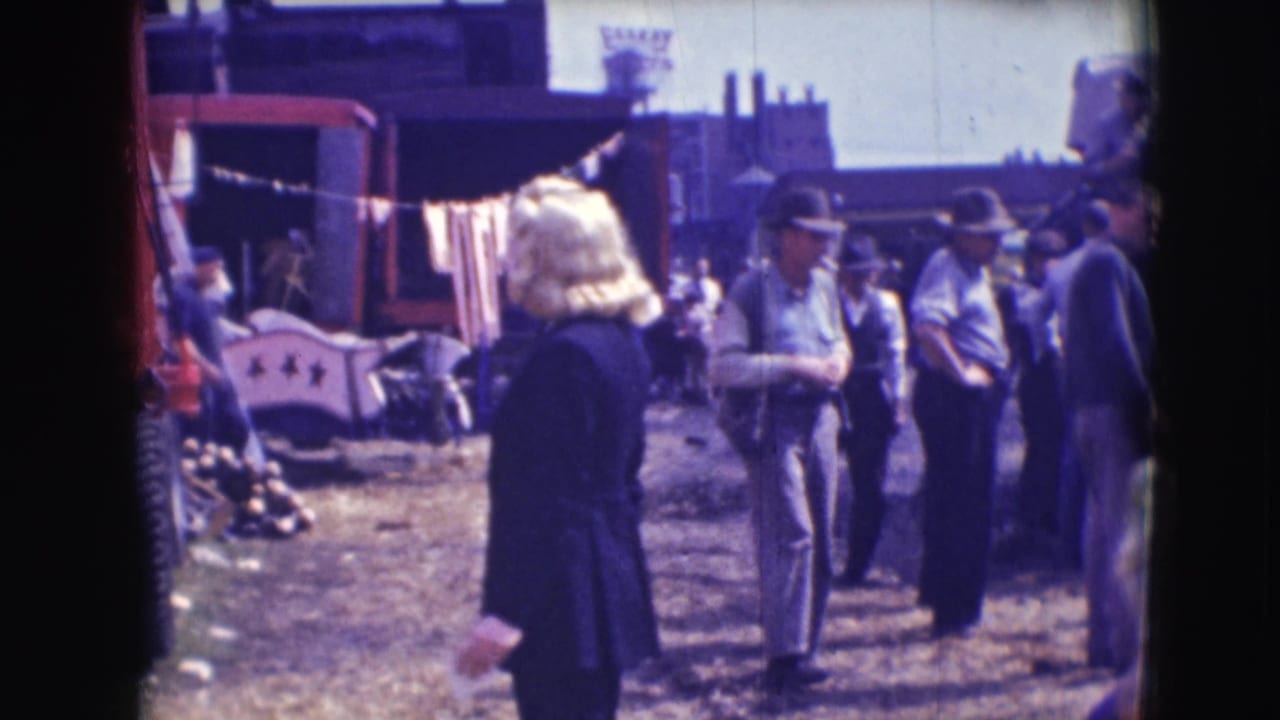 Circus grounds with a red truck, laundry line, workers, and a blonde person in the foreground.