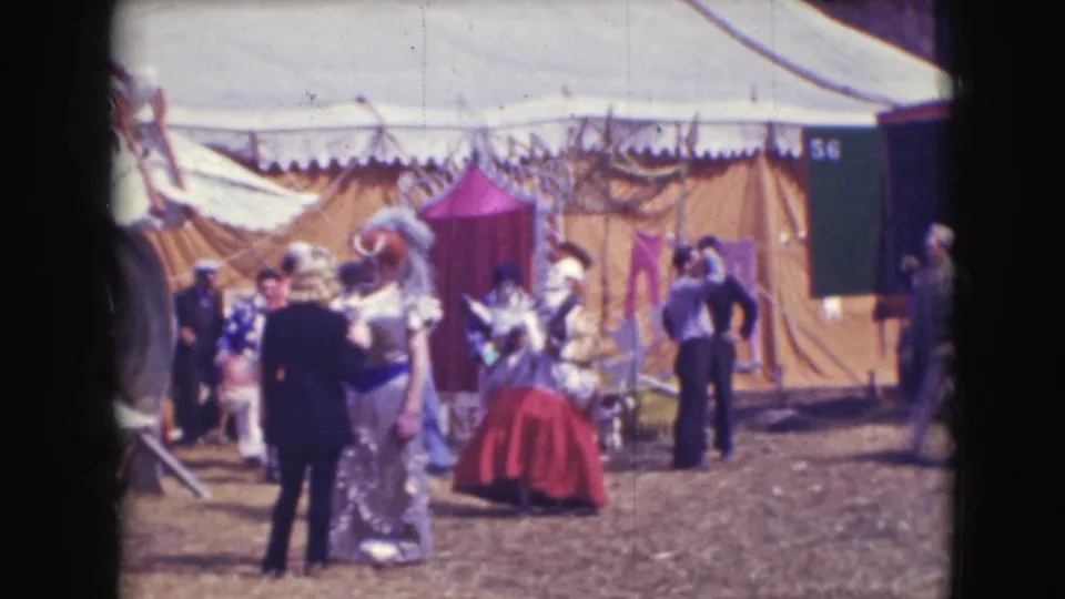 A costumed clown in a red skirt stands near a yellow circus tent in the Ringling Brothers Sarasota setting, with performers and staff around them.