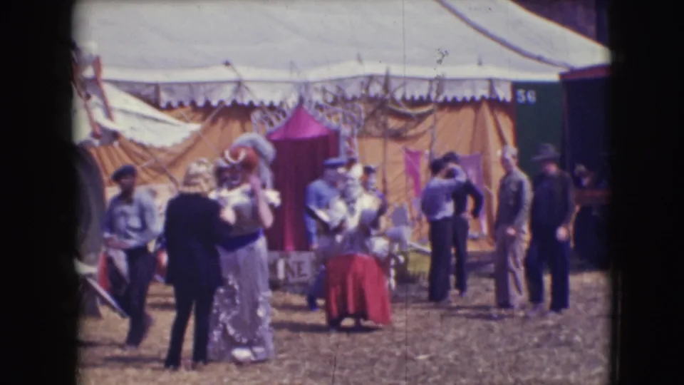 Outside the Sarasota circus tent, the red-skirted clown faces away while staff and costumed performers stand nearby on the dirt grounds.