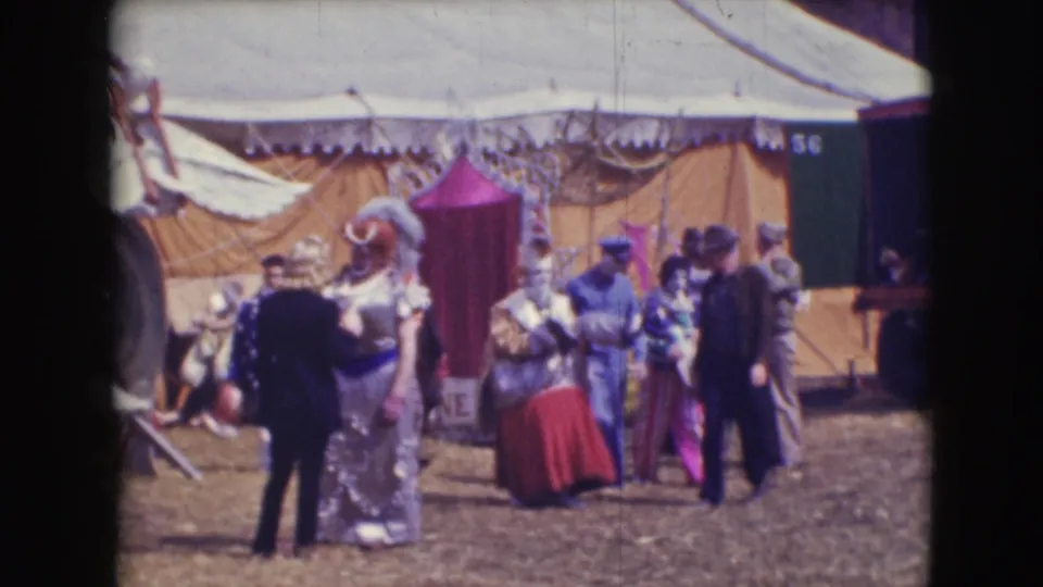 The red-skirted clown walks away across the circus grounds near the yellow tent, with men and other performers clustered in the background.