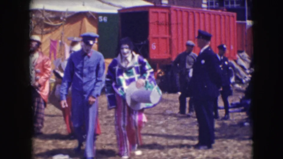 A checkered-costume clown walks through the 1948 Sarasota circus lot with uniformed staff, tents, and a red rail car behind.