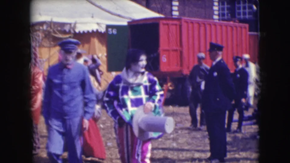 In the Sarasota circus lot, a uniformed man walks beside a clown in a purple and green checkered costume near tents and a red rail car.
