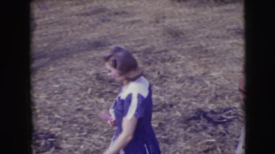 Side view of a woman in a purple and white dress walking through the 1948 Sarasota circus-event grounds on 8mm film.