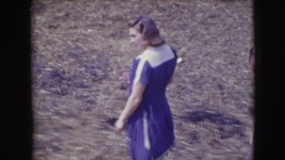 Rear three-quarter view of the woman in a purple and white dress moving through the Sarasota circus-event grounds in 1948 footage.