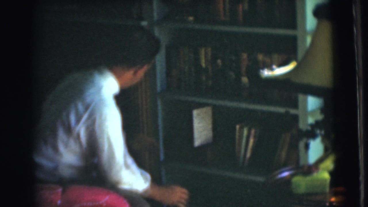 Man in a white shirt sits beside dark bookshelves in a dim study.