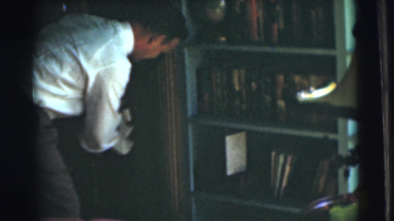 Man bends forward near bookshelves with a lamp visible at the side.