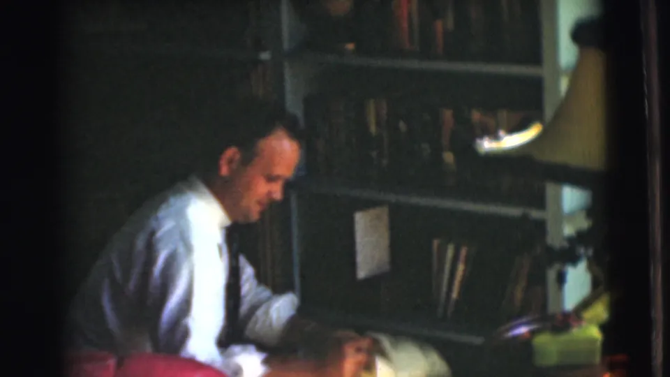A seated man in a white shirt and tie appears to handle a book or papers beside shelves in the supplied 1951 Chatham interior footage.