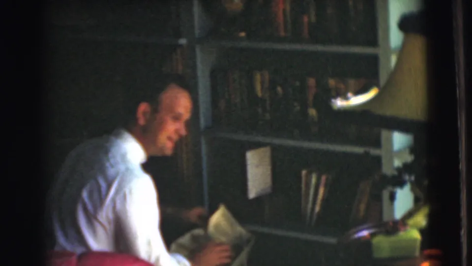 In a dim private study from the supplied 1951 Chatham home-movie context, the man smiles while holding papers or a book near the shelves.