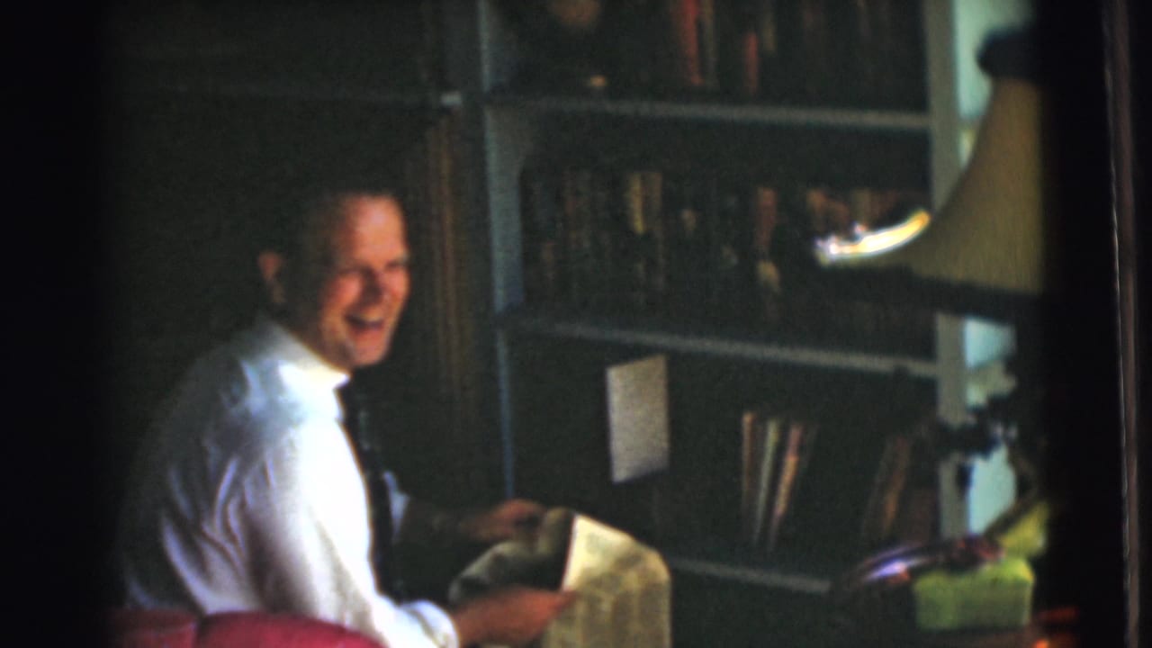 Man in a white shirt and tie sits by a bookcase holding a newspaper.
