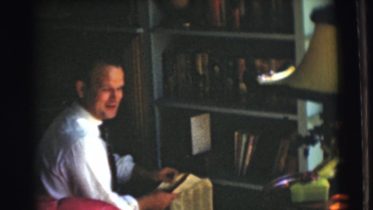 The man sits in the Chatham home interior with a newspaper in hand, a bookcase and shaded lamp behind him.