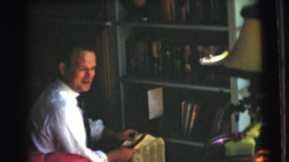 The man sits in the Chatham home interior with a newspaper in hand, a bookcase and shaded lamp behind him.