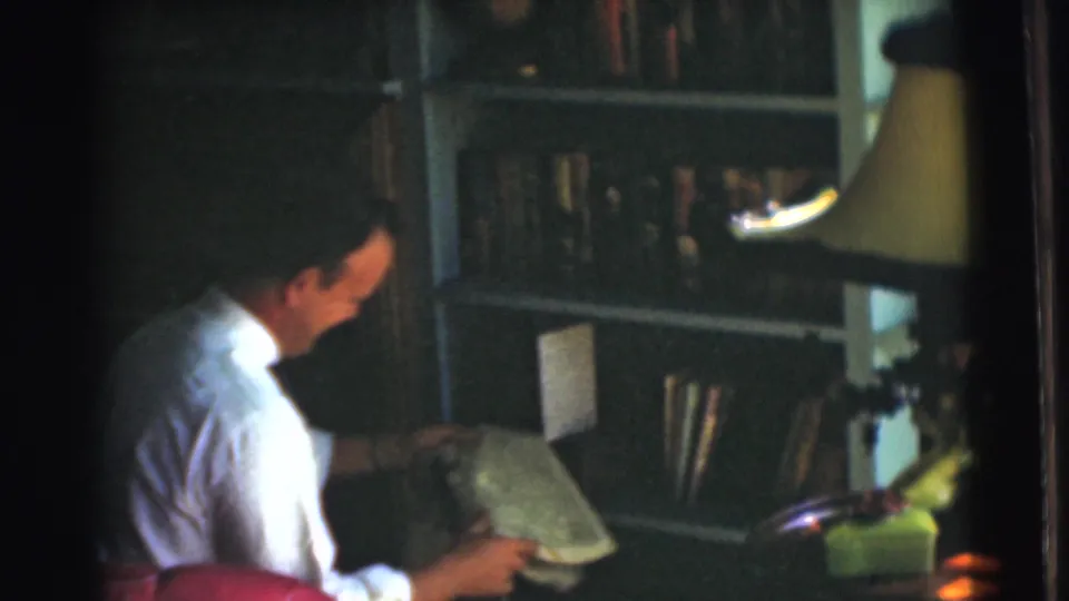 The seated man lowers his gaze to an open newspaper, with bookshelves and a lamp visible in the 1951 home interior.