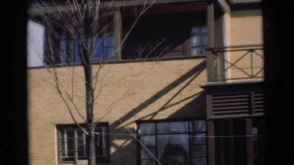1951 Chatham, New Jersey 8mm frame showing a tan brick building facade with balcony railings, windows, tree branches, and strong shadows.