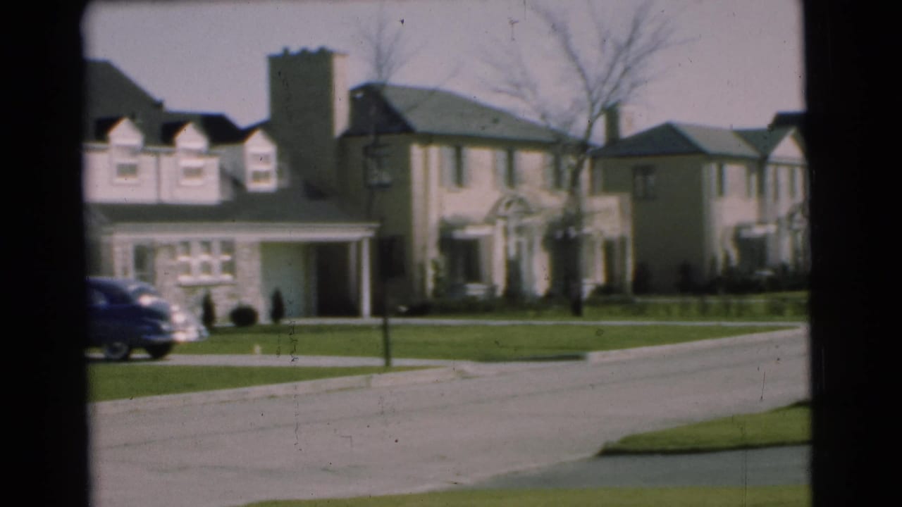 1951 Chatham, New Jersey home-movie frame showing a suburban street with two-story houses, green lawns, chimneys, and a sedan passing at left.