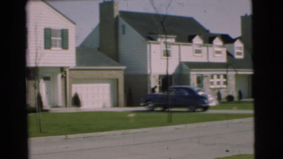 1951 Chatham, New Jersey home-movie frame showing closely spaced two-story homes, garage doors, lawns, young trees, and a dark sedan.