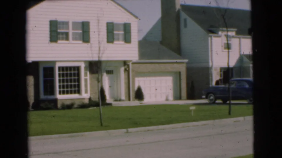 1951 Chatham, New Jersey frame showing neighboring two-story houses with shutters, garage doors, lawns, a young tree, and a sedan at right.