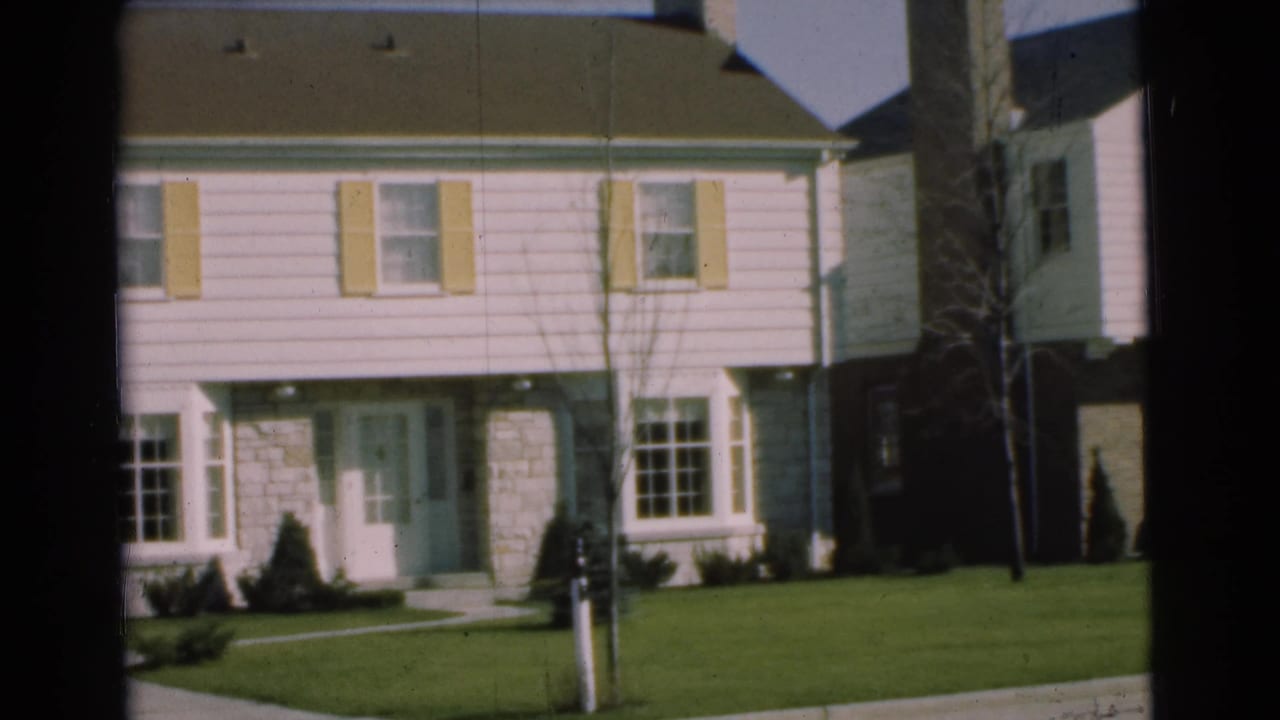 1951 Chatham, New Jersey home-movie frame showing a closer view of a two-story house front with shutters, doorway, lawn, and sidewalk.