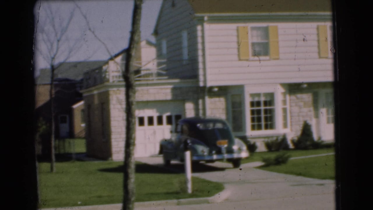 Suburban house, driveway, lawn, tree trunk, and parked dark car near the garage.