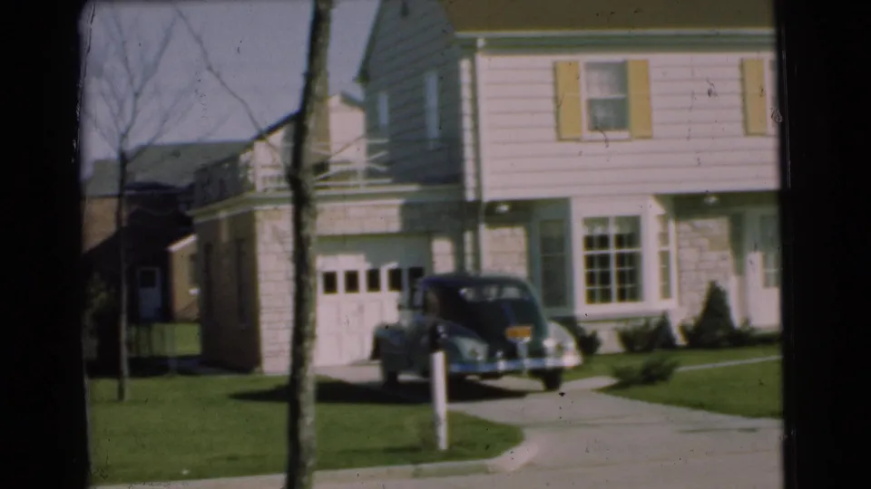 1951 Chatham residential exterior with a dark sedan parked by a light-sided house and garage, seen across a green lawn.