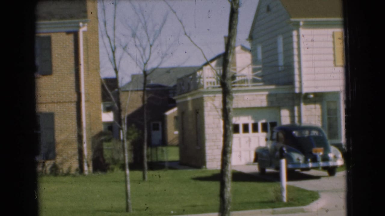 1951 Chatham home-movie frame panning past neighboring suburban houses, a parked sedan, sidewalk, lawns, and bare trees.
