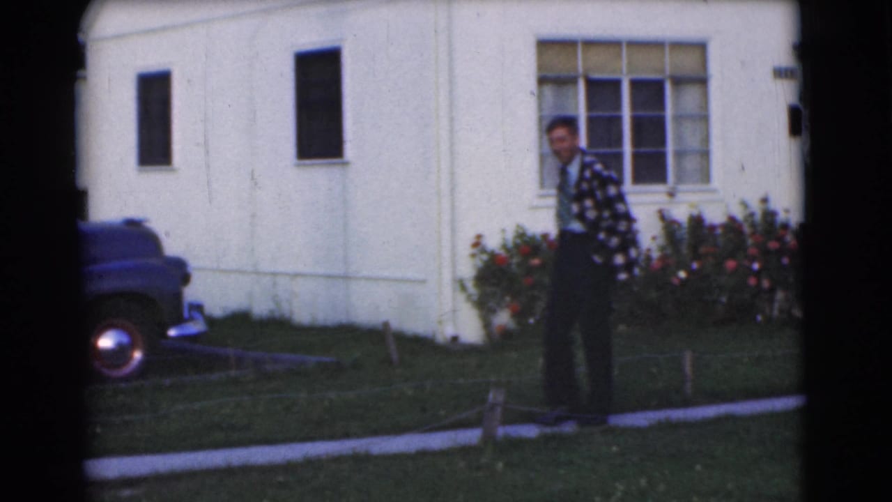 Young man in a plaid jacket standing near a sidewalk with a blue car and pale house behind him.