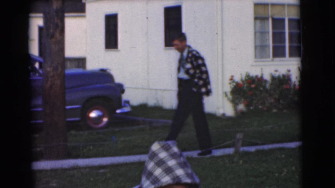 The young man walks along a suburban sidewalk in the supplied Chatham, New Jersey 1951 scene, passing a parked blue car and house exterior.