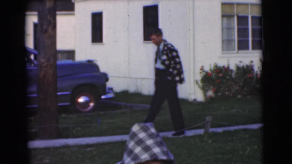 The young man walks along a suburban sidewalk in the supplied Chatham, New Jersey 1951 scene, passing a parked blue car and house exterior.