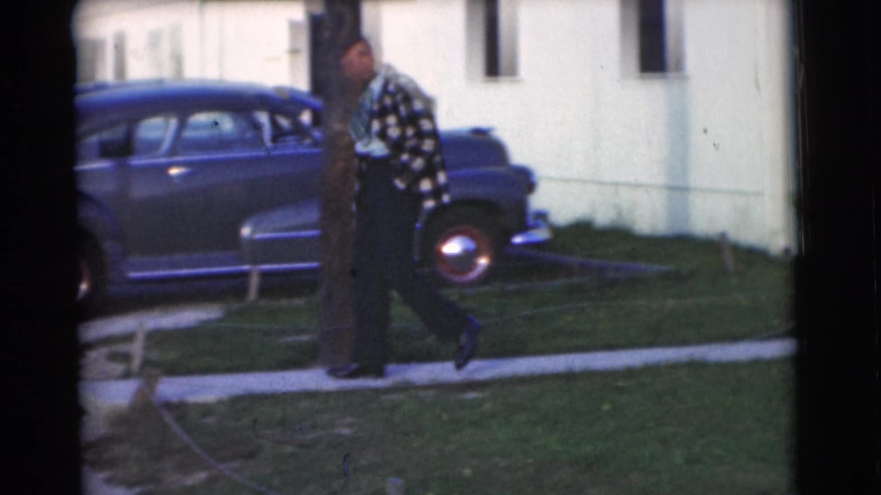 The man continues past a rounded blue sedan in the supplied 1951 Chatham home-movie footage, with lawn and house exterior visible.