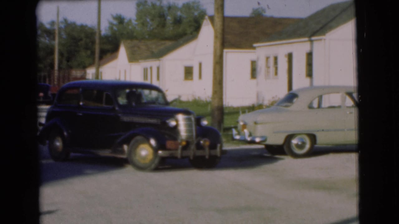 Black sedan moving past a utility pole with a pale car parked nearby.